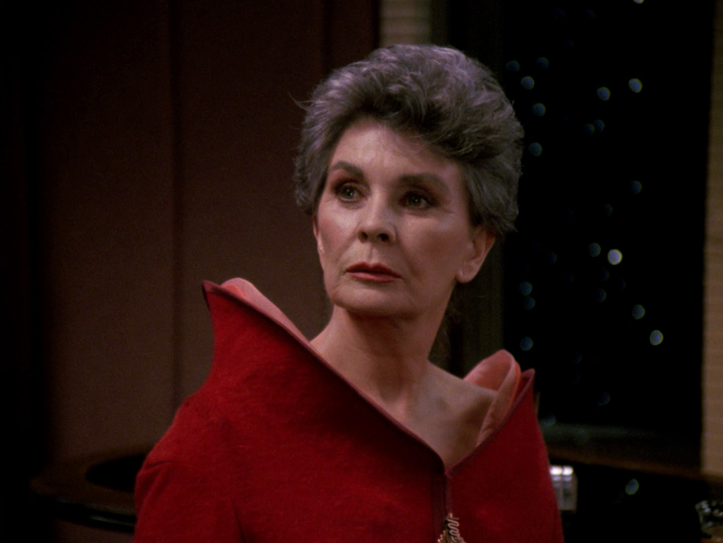 Screencap of Jean Simmons as Norah Satie: an elegant woman in her early 60s with short, dark grey hair, wearing a red gown with an asymmetric vertical collar. 